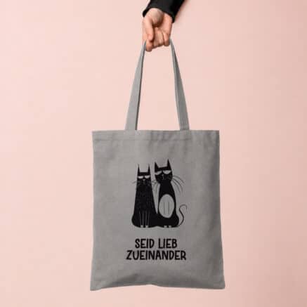 Grauer Baumwollbeutel mit zwei schwarzen Katzen und dem Spruch „Seid lieb zueinander“, gehalten vor einem zartrosa Hintergrund.