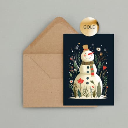 Weihnachtskarte mit fröhlichem Schneemann, Zylinder und Schal auf dunkelblauem Hintergrund. Winterliche Blumen, Zweige und Schneeflocken sowie goldene Veredelung schmücken das Motiv. Die Karte liegt vor einem braunen Kraftpapier Umschlag.